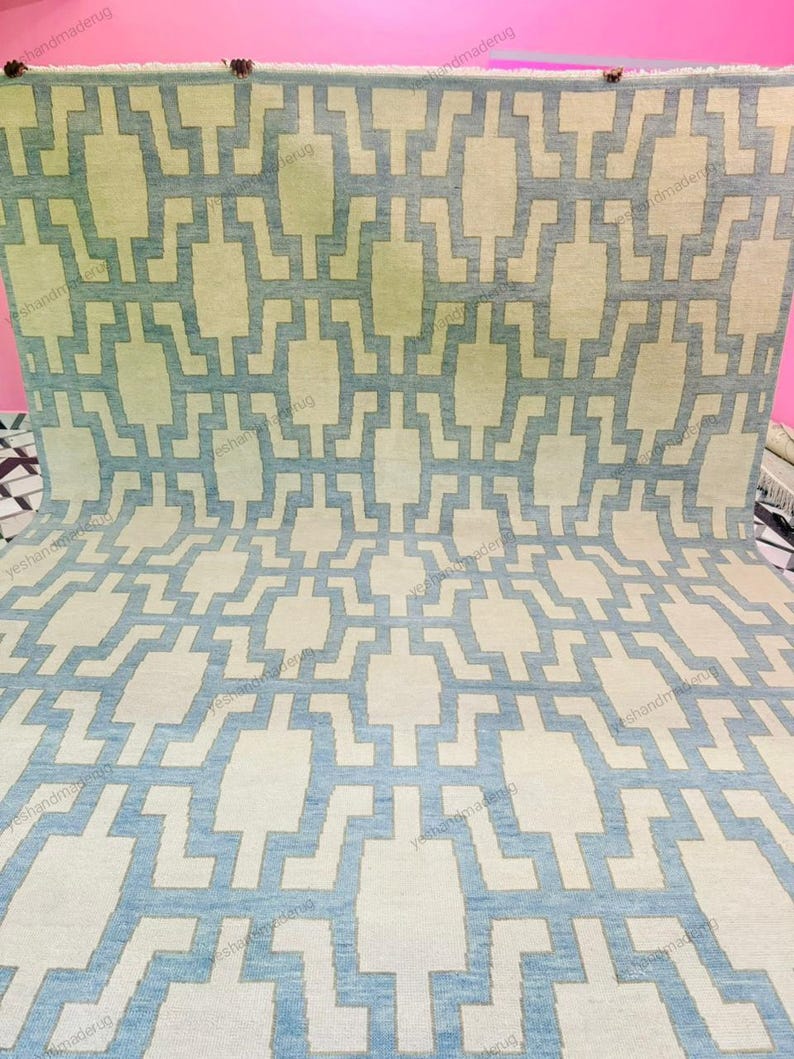 Room scene featuring a large handmade wool rug placed on a patterned floor, showcasing its modern geometric design. The calming blue and beige tones enhance the overall interior aesthetic. This flatweave area rug is ideal for contemporary homes, offering durability, comfort, and style. Perfect for living rooms or bedrooms, it blends handcrafted quality with modern home décor trends.