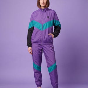 May include: A purple tracksuit featuring a zip-up jacket and matching pants. The jacket has a high collar, black sleeves, and a teal chevron stripe across the chest. The pants have teal chevron stripes on the legs and elastic cuffs.