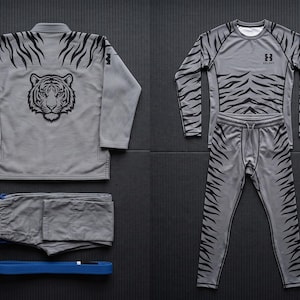 May include: A grey martial arts uniform set featuring a jacket with a tiger head graphic on the back, matching folded pants, a blue belt, and a long-sleeve compression shirt and leggings set, both decorated with a black tiger stripe pattern.