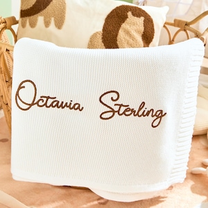 Puede incluir: Una manta blanca para bebé con el nombre "Octavia Sterling" bordado en marrón. La manta está doblada y colocada en una cesta de mimbre, junto con un cordero de peluche y una almohada con un diseño de león.