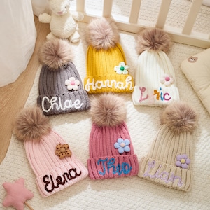 Puede incluir: Varios gorros de punto personalizados en colores como gris, amarillo, rosa y crema. Cada gorro tiene un pompón, un adorno floral y un nombre bordado en un color contrastante. Los nombres incluyen Chloe, Hannah, Eric, Elena, Iris y Lilian.