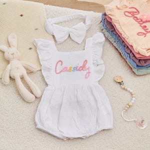 Puede incluir: Un pelele blanco para bebé con el nombre "Cassidy" bordado en colores arcoíris, una diadema a juego con un lazo y un juguete de peluche de conejito. También hay un chupete con una cadena de cuentas de madera.