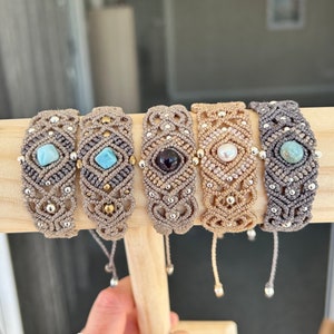 May include: Five macrame bracelets with different coloured gemstones. The bracelets are made with brown cord and silver beads. The gemstones are turquoise, amethyst, pearl, and white.