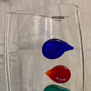 May include: A clear glass vase with a textured surface and a label that reads "Barbinini Murano". The vase is decorated with seven colorful glass drops in shades of blue, red, green, and yellow.