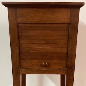 May include: A wooden cabinet with a shuttered door and a single drawer. The cabinet is brown and has a simple design. It is standing on a tiled floor.