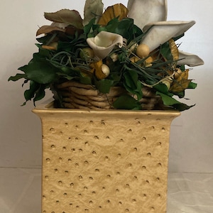 Puede incluir: Un macetero cuadrado de cerámica beige con una superficie texturizada. El macetero tiene una cesta tejida en el interior con un arreglo floral de flores artificiales blancas, marrones y verdes.