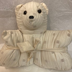 Op de afbeelding: Een witte knuffelbeer met een crèmekleurig stoffen lichaam en zwarte ogen. De beer is vastgebonden met een crèmekleurig lint en heeft een crèmekleurig stoffen patroon met kleine vierkantjes.