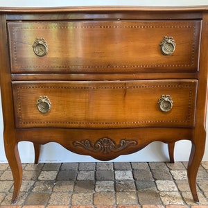 Può includere: Un comò in legno a due cassetti con un fronte curvo e intagli decorativi. Il comò ha una finitura in legno marrone chiaro e tiretti in ottone.