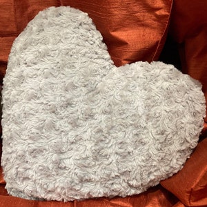 May include: A heart-shaped decorative pillow in a soft, light gray color. The pillow has a textured surface with a rose-like pattern, giving it a plush appearance. The pillow is resting on a reddish-orange fabric.