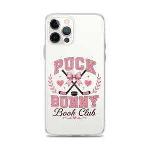 May include: Clear phone case with a pink and white design. The design includes the text "PUCK BUNNY Book Club" with crossed hockey sticks, a pink bow, hearts, and a laurel wreath. The case is designed to fit a smartphone.
