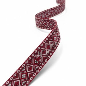 May include: A long, narrow strip of woven fabric featuring a repeating geometric pattern in deep red, white, and black. The design consists of interconnected diamond shapes and small cross motifs arranged in a symmetrical, linear sequence along the ribbon.