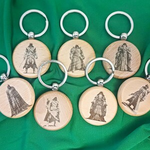 Può includere: Una collezione di portachiavi in legno con anelli argentati. Ogni portachiavi presenta un'immagine incisa diversa di un personaggio. I portachiavi sono rotondi e realizzati in legno chiaro. Lo sfondo è un tessuto verde unito.