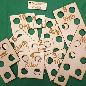 Puede incluir: Piezas de madera de Numicon con números y palabras galesas grabadas. Las piezas tienen varias formas y tamaños, con recortes circulares. El texto "Numicon Welsh" está en una pequeña pieza rectangular. Sobre una superficie verde.