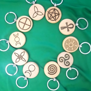 Może przedstawiać: Kolekcja drewnianych breloków do kluczy z różnymi wygrawerowanymi symbolami, w tym pentagramem i ankh. Każdy brelok ma srebrny pierścień. Breloki są ułożone na zielonym tle.