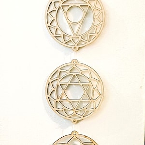 Może przedstawiać: Siedem drewnianych wiszących ozdób ściennych, każda z innym geometrycznym wzorem, reprezentującym siedem czakr. Wzory są skomplikowane i szczegółowe, a drewno jest jasnobrązowe.