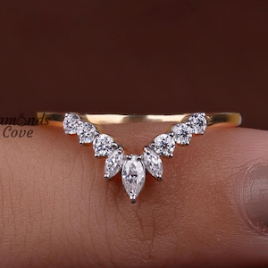 Könnte beinhalten: Ein goldfarbener Ring mit geschwungenem Design, besetzt mit mehreren kleinen, runden und marquiseförmigen, klaren Edelsteinen. Der Ring wird von einem Finger vor braunem Hintergrund gehalten. Der Text "Diamonds Cove" ist sichtbar.