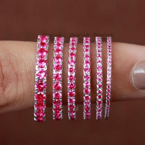 Può includere: Un set di sette sottili fedi nuziali in argento con pietre preziose rosa. Le fedi sono impilate su un dito, con la fede più piccola in basso e la fede più grande in alto.