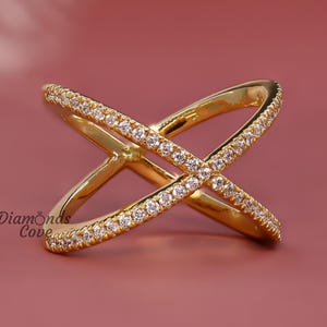 May include: A gold-colored criss-cross ring with small, clear gemstones set along the top of each band. The ring is displayed on a pink background. The ring is a piece of jewelry.