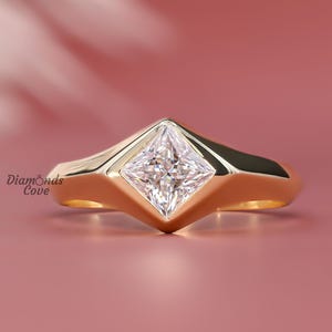 May include: A gold ring with a square-cut diamond center stone. The ring has a unique, geometric design with a wide band and a bezel setting. The ring is displayed on a pink background, with the text "Diamonds Cove" visible.