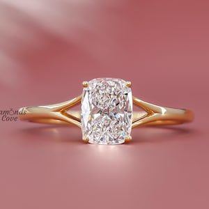 Puede incluir: Un anillo de oro con una gema grande, rectangular y transparente. El anillo tiene un diseño de caña dividida. El fondo es de color rosa apagado sólido. El texto "Diamonds Cove" está en la esquina inferior izquierda.