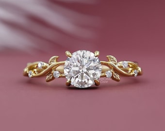 May include: A gold engagement ring with a round diamond center stone and a delicate vine design. The ring is set on a pink background.