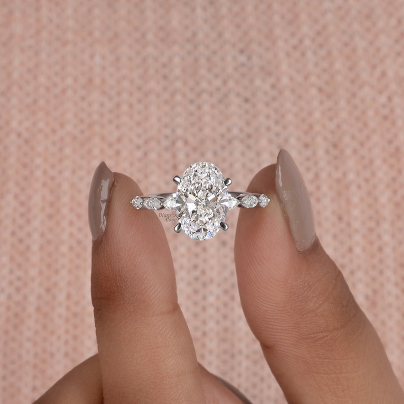 May include: A silver engagement ring with an oval-shaped diamond center stone and smaller diamond accents on the band. The ring is on a finger.