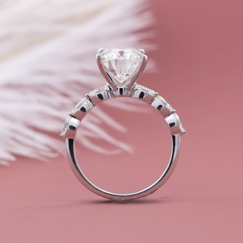 May include: A silver engagement ring with a large round center diamond and smaller round diamonds set in a pave band. The ring is on a white feather against a pink background.