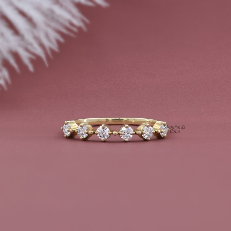 May include: A delicate gold band ring with a row of small, round diamonds set in a staggered pattern. The ring is on a pink background.