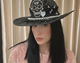Puede incluir: Sombrero de paja negro con ala ancha, adornado con un diseño de calavera y huesos cruzados. El sombrero presenta una banda decorativa con un motivo de calavera y una pluma. Está adornado con pedrería y detalles blancos.