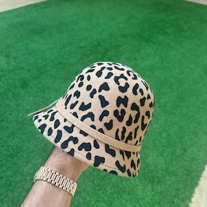 May include: A tan cloche hat with a black leopard print design. The hat features a tan band and a small brim. The hat is held up by a person's hand.