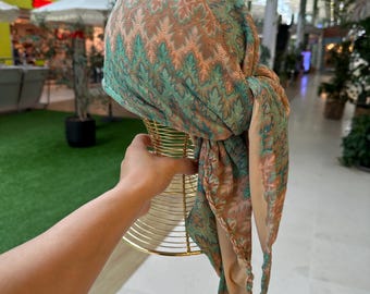 May include: A patterned head covering in shades of teal, peach, and brown is draped over a gold wire form. The fabric has a textured design and long, flowing ends. The head covering is likely a turban or scarf.