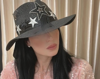 Puede incluir: Sombrero de paja negro con ala ancha, adornado con aplicaciones de estrellas blancas y un diseño central de estrella con pedrería. El sombrero tiene una cinta negra y flecos.
