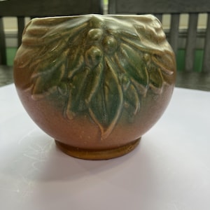 May include: A ceramic vase with a round body and a wide opening. The vase features a design of leaves and berries in shades of green and brown. The base of the vase is a darker brown.