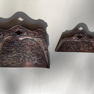 May include: Two antique copper-colored metal dustpans. The larger dustpan has a curved top with a diamond-shaped cutout and is decorated with floral patterns. The smaller dustpan has a similar design. Both are placed on a white surface.