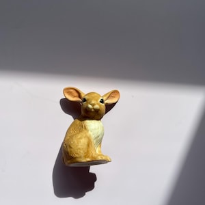 May include: A ceramic figurine of a rabbit in a seated position. The rabbit has large, upright ears and a light brown coat with white accents. The figurine is on a white surface, with shadows cast to the side.