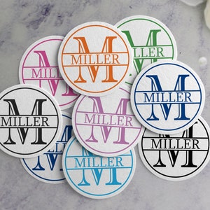 May include: A set of eight round coasters with a white background and a black, blue, green, orange, pink, or purple circle in the center. Each circle features a large letter "M" with the word "MILLER" printed in a smaller font below the letter.