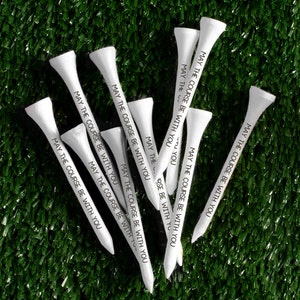 May include: A set of white golf tees with the phrase "May the course be with you" printed on them. The tees are arranged in a fan shape on a green artificial grass surface.