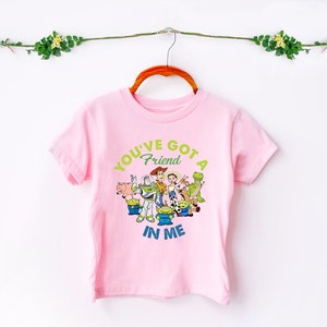 May include: Pink short-sleeved t-shirt featuring a graphic of the Toy Story characters. The text on the shirt reads "You've got a friend in me".