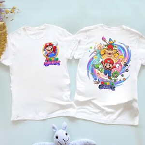 Peut inclure: Deux t-shirts blancs avec des graphiques du jeu vid&eacute;o "The Super Mario Galaxy". Un t-shirt a un petit graphisme de Mario sur le devant. L'autre t-shirt a un graphisme plus grand de Mario et d'autres personnages au dos.