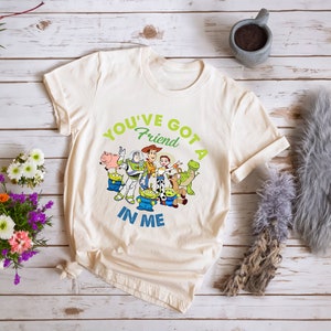 May include: A beige t-shirt featuring a green and blue graphic of the Toy Story characters Woody, Buzz Lightyear, and others. The text "You've Got A Friend In Me" is printed on the shirt.