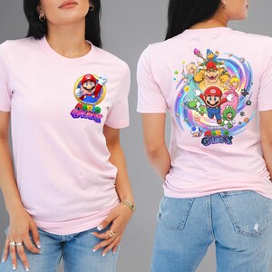 Peut inclure: T-shirt rose clair avec un graphisme color&eacute; de Mario du jeu vid&eacute;o Super Mario Galaxy. Le devant pr&eacute;sente un motif circulaire de Mario, tandis que le dos affiche un motif plus grand avec plusieurs personnages et le logo du jeu.