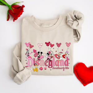 Puede incluir: Una sudadera beige con un gr&aacute;fico rosa y rojo de Mickey Mouse, Minnie Mouse, Donald Duck y Daisy Duck sosteniendo globos en forma de coraz&oacute;n. El gr&aacute;fico dice "Disneyland Sweethearts Nite".