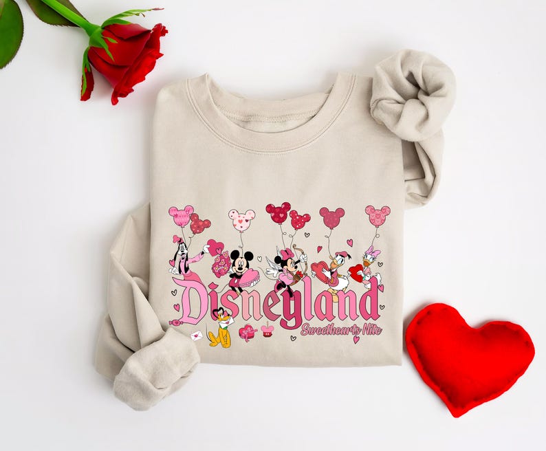 Puede incluir: Una sudadera beige con un gr&aacute;fico rosa y rojo de Mickey Mouse, Minnie Mouse, Donald Duck y Daisy Duck sosteniendo globos en forma de coraz&oacute;n. El gr&aacute;fico dice "Disneyland Sweethearts Nite".