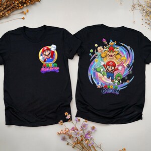 Peut inclure: Deux t-shirts noirs avec des graphiques du jeu vid&eacute;o Super Mario Galaxy. Un t-shirt a un graphisme avant de Mario dans un cercle dor&eacute; avec le titre du jeu. L'autre t-shirt a un grand graphisme de Mario et d'autres personnages.