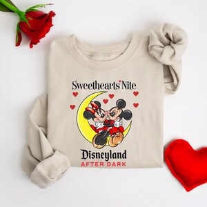 Puede incluir: Una sudadera beige con un gr&aacute;fico de Mickey y Minnie Mouse bes&aacute;ndose en una luna creciente. El texto "Sweethearts Nite Disneyland After Dark" est&aacute; impreso debajo del gr&aacute;fico.