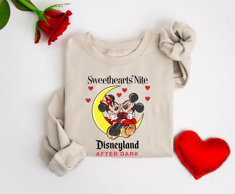 Puede incluir: Una sudadera beige con un gr&aacute;fico de Mickey y Minnie Mouse bes&aacute;ndose en una luna creciente. El texto "Sweethearts Nite Disneyland After Dark" est&aacute; impreso debajo del gr&aacute;fico.