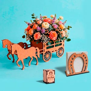 Puede incluir: Un carro decorativo de madera lleno de flores coloridas, tirado por dos recortes de caballos de madera. Un servilletero de herradura de madera y una pequeña caja con texto también están presentes, todo sobre un fondo azul.