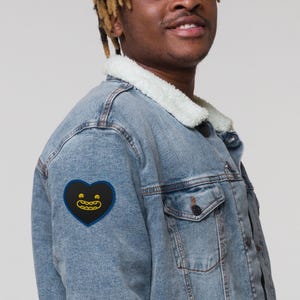 May include: A light blue denim jacket with a white sherpa collar. A black heart-shaped patch with a yellow smiling face is sewn on the left side of the jacket. The jacket has two chest pockets and a button closure.