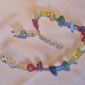 May include: A rainbow colored beaded bracelet with a silver clasp. The bracelet features small, irregular shaped gemstones in shades of green, blue, purple, red, and yellow.