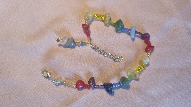 May include: A rainbow colored beaded bracelet with a silver clasp. The bracelet features small, irregular shaped gemstones in shades of green, blue, purple, red, and yellow.